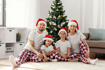 family, winter holidays and people concept - happy mother, father and two daughters in santa hats sitting under christmas tree at home