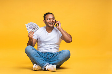Portrait of african american indian black young man sitting on the floor holding dollar banknotes isolated over yellow background. Using mobile phone.