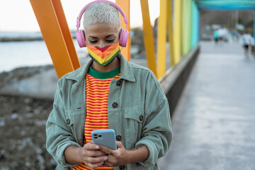 Young gay woman wearing LGBT face mask and using mobile smartphone while listening to music with...
