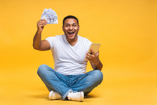 Portrait Of African American Indian Black Young Man Sitting On The Floor Holding Dollar Banknotes Isolated Over Yellow Background. Using Tablet Computer.