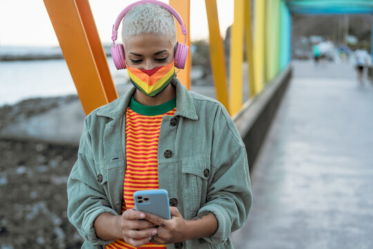 Young gay woman wearing LGBT face mask and using mobile smartphone while listening to music with wireless headphones outdoor - Gender equality and technology concept