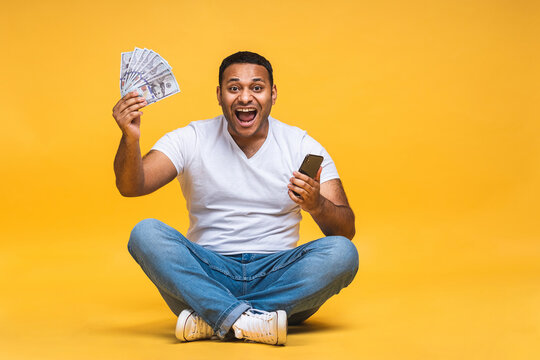 Portrait of african american indian black young man sitting on the floor holding dollar banknotes isolated over yellow background. Using mobile phone.