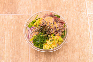 Poke bowl in plastic takeaway container with red onion, wakame seaweed, beef, ripe avocado and spinach