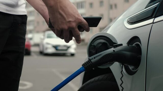 Unrecognizable man disconnects charging cable to to electric vehicle. Male hand unplugs power connector into EV car and charges batteries, uses phone to disconnect charging