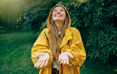 Image of a positive young blonde woman smiling wearing yellow raincoat during the rain in the park....