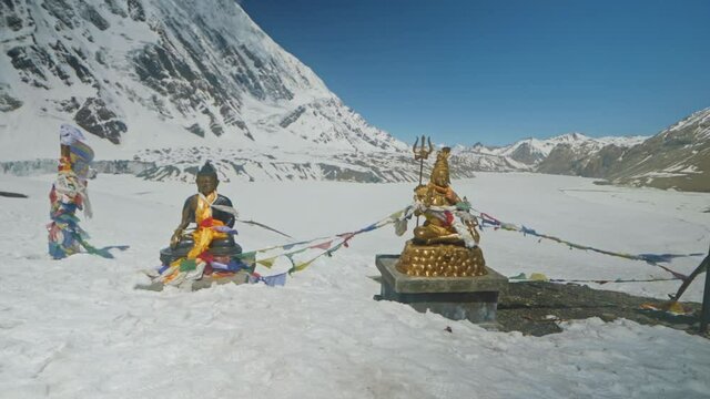 Statues of Bouddha and Lord Shiva shine at sun against frozen highland Tilicho Lake at foot of snowy Tilicho Peak, Nepal