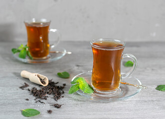 Two Turkish cups of tea stand on a light wooden table. Mint leaves and tea leaves are scattered in...