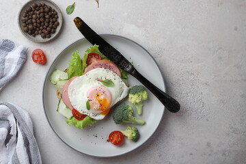 beautiful healthy food healthy breakfast sandwich with poached egg, lettuce, broccoli and cherry tomatoes on a plate on a gray background under concrete top view, with a knife and a napkin modern styl