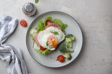 beautiful healthy food healthy breakfast sandwich with poached egg, lettuce, broccoli and cherry tomatoes on a plate on a gray background under concrete top view, with a knife and a napkin modern styl