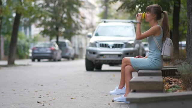 Young woman drinking coffee from paper cup sitting on city street bench in summer park.

