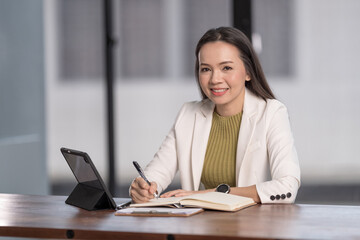 Adult cheerful businesswoman in business outfits writing on notebook with digital table at her working desk with in office