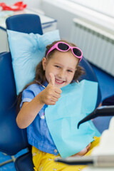 little girl is sitting in a chair at a dentist's appointment. The child shows a sign of approval, a thumbs up after professional treatment in children's dental polyclinic. treat baby teeth with milk.