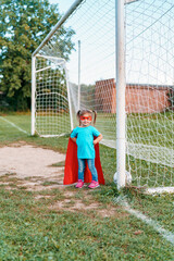 little girl child in a superhero costume is playing football in the fresh air. happy girl in red raincoat and a mask runs with a ball on the football field. Outdoor games, dynamic image
