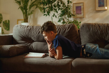 Fototapeta na wymiar An attentive boy is reading a book lying on the couch at home. The student is passionate about reading. The child prepares homework, does homework lying down.