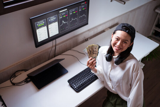 Smiling Asian trader cooling down with paper dollars against monitor