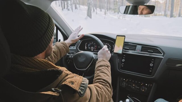 Handheld shot of man in warm winter clothes driving car along narrow forest road covered in snow. His girlfriend sitting on passenger seat beside him