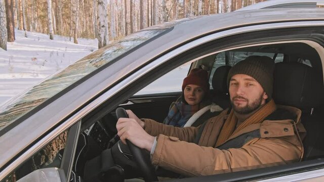 PAN portrait of couple in warm hats and parkas posing for camera in car parked in forest area in winter