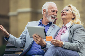 Senior couple sitting in the park on bench and shopping on line with tablet and credit card