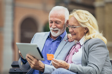 Senior couple sitting in the park on bench and shopping on line with tablet and credit card