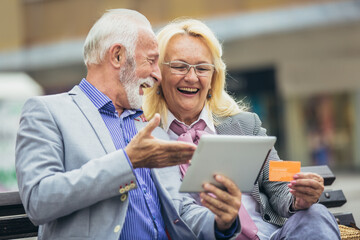 Senior couple sitting in the park on bench and shopping on line with tablet and credit card