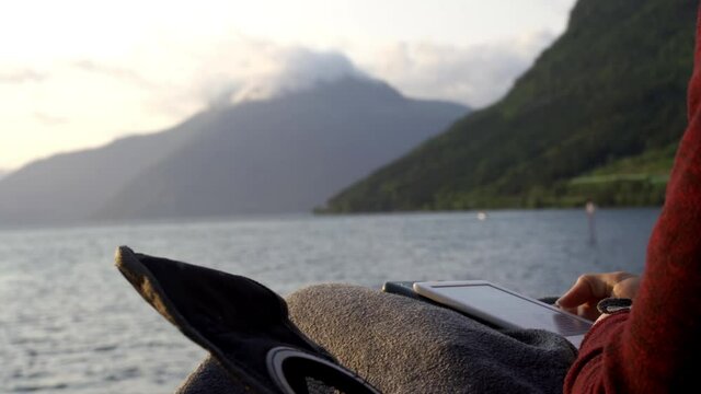 Hiker Scrolling Tablet In Chair On Riverbank At Sunset