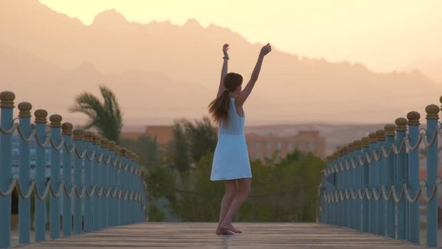 Young happy woman in light dress dancing on long pier at seaside with silhouette of mountain peaks in the evening. Vacation and traveling concept.