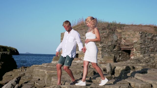 Young couple expecting a baby is walking on big stones at the seaside. Bright sunny summer day and a couple walking on stones to enjoy marine landscape together