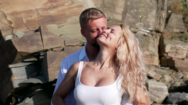A man  is embracing his pregnant wife, rubbing her belly and kissing her with love and tenderness against rocky cliff. Woman enjoys the sun and his husband