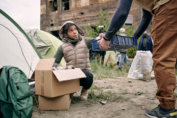 Dark-skinned teenage girl crouching at stuff box and putting tin can into basket while refugees...