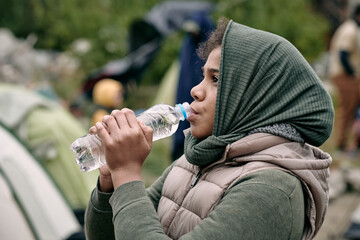 Teenage middle-eastern girl with scarf covering head drinking water greedily in refugee camp
