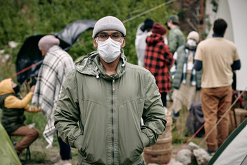 Portrait of middle-eastern man in facial mask holding hands in jackets pockets and standing against crowd of migrants