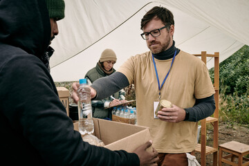 Young bearded volunteer in glasses holding tin can and giving bottle of water to homeless man with...