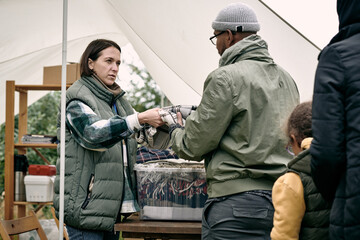 Female social worker in vest standing under tent and giving plaid to refugee while providing it to them