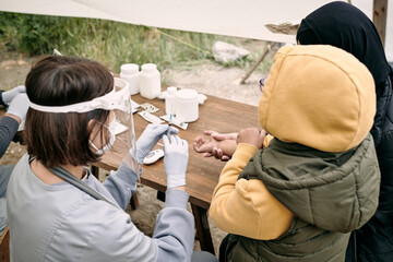 Nurse in gloves and mask preparing syringe while vaccinating refugee child outdoors