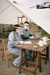 Team of doctors in safety face shields sitting at table in tent and working with medical cards of...