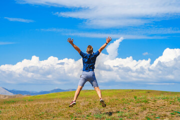 Rear view of a happy young man jumping on a summer meadow in the air against a blue sky with clouds
