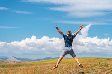 Rear view of a happy young man jumping on a summer meadow in the air against a blue sky with clouds