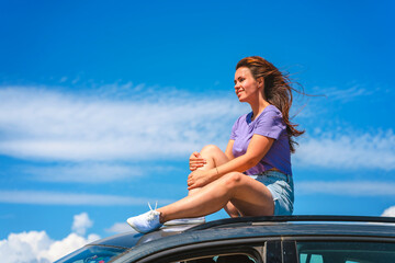 Fototapeta na wymiar A young woman in shorts is sitting on the roof of a Volkswagen Tiguan car with a view of a beautiful blue sky with clouds. The concept of traveling by car. Sudak, Crimea - 2 Sep 2021