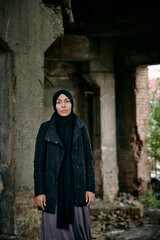 Portrait of sad refugee young Muslim woman in hijab and long jacket standing against ruined building