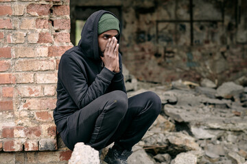 Distraught young Black refugee man in black outfit crouching at ruined building and covering face...