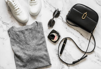 Women's clothing set - cashmere gray pullover, cross body black leather bag, sunglasses, cosmetics, white sneakers on a marble background, top view