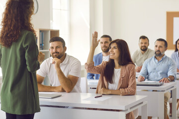 Smiling young woman from student audience raises hand to ask teacher a question. Group of adult people listening to business trainer sitting at tables in classroom during corporate training or seminar