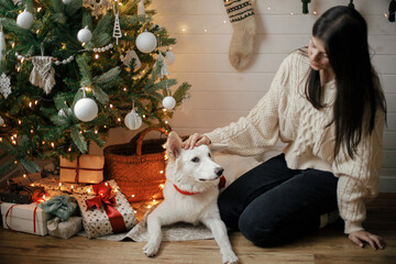 Stylish happy woman in cozy sweater sitting with adorable dog under christmas tree with gifts and lights. Young female caressing cute dog in festive scandinavian room. Pet and winter holidays