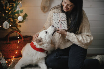 Merry Christmas! Stylish happy woman with christmas gift and adorable dog sitting under christmas tree with lights. Cute dog and young female with wrapped present sitting in festive scandinavian room