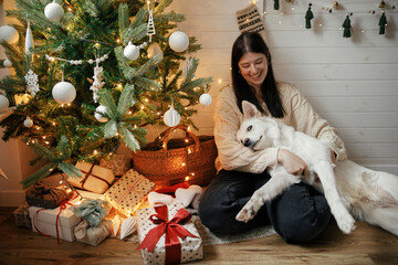 Stylish happy woman sitting and hugging adorable dog under christmas tree with gifts and lights....