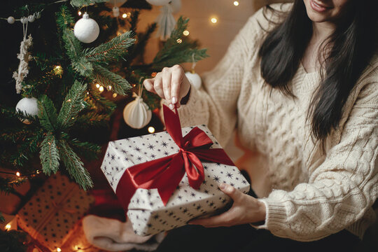 Hands In Cozy Sweater Opening Christmas Gift With Red Bow On Background Of Christmas Tree With Lights. Stylish Female Holding Present With Red Ribbon In Festive Room Close Up. Merry Christmas!