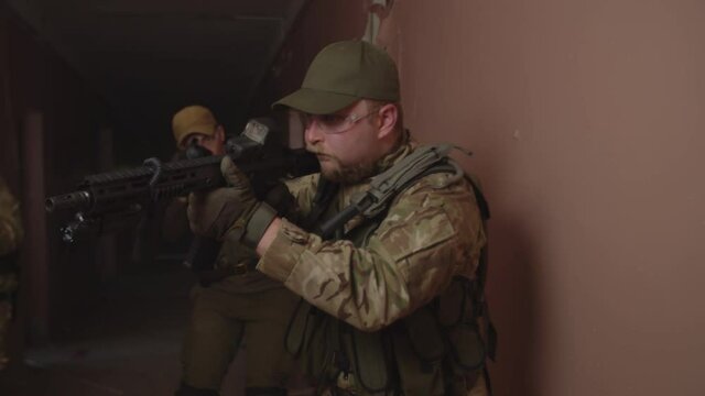 Portrait of fully equipped stormtrooper in combat gear, assault rifle in firing position, checking room while anti-terrorist group of soldiers walking along dirty corridor of abandoned enemy building