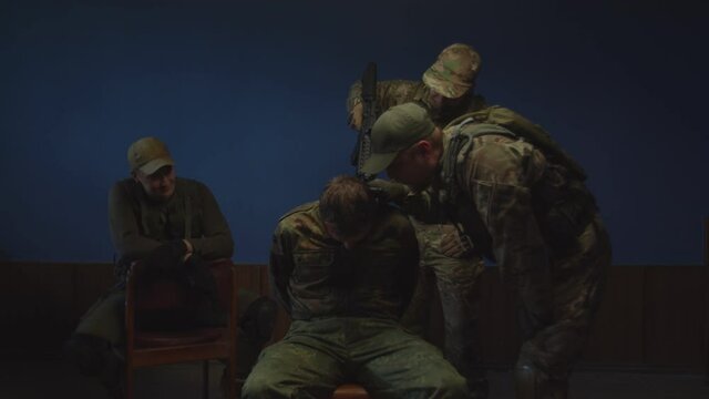 Brutal serious soldier in combat gear with gun intimidating male terrorist indoor, prying out information while second military man putting weapon to prisoner head during interrogation indoors.