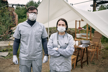 Portrait of young medical workers in masks and scrubs standing against tent with medicine in...