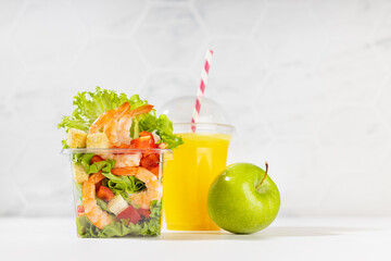 Wellness fresh take away food - set of orange juice, green apple and  tropical salad with shrimps, lettuce, red bell pepper, croutons in plastic box in white bar interior.
